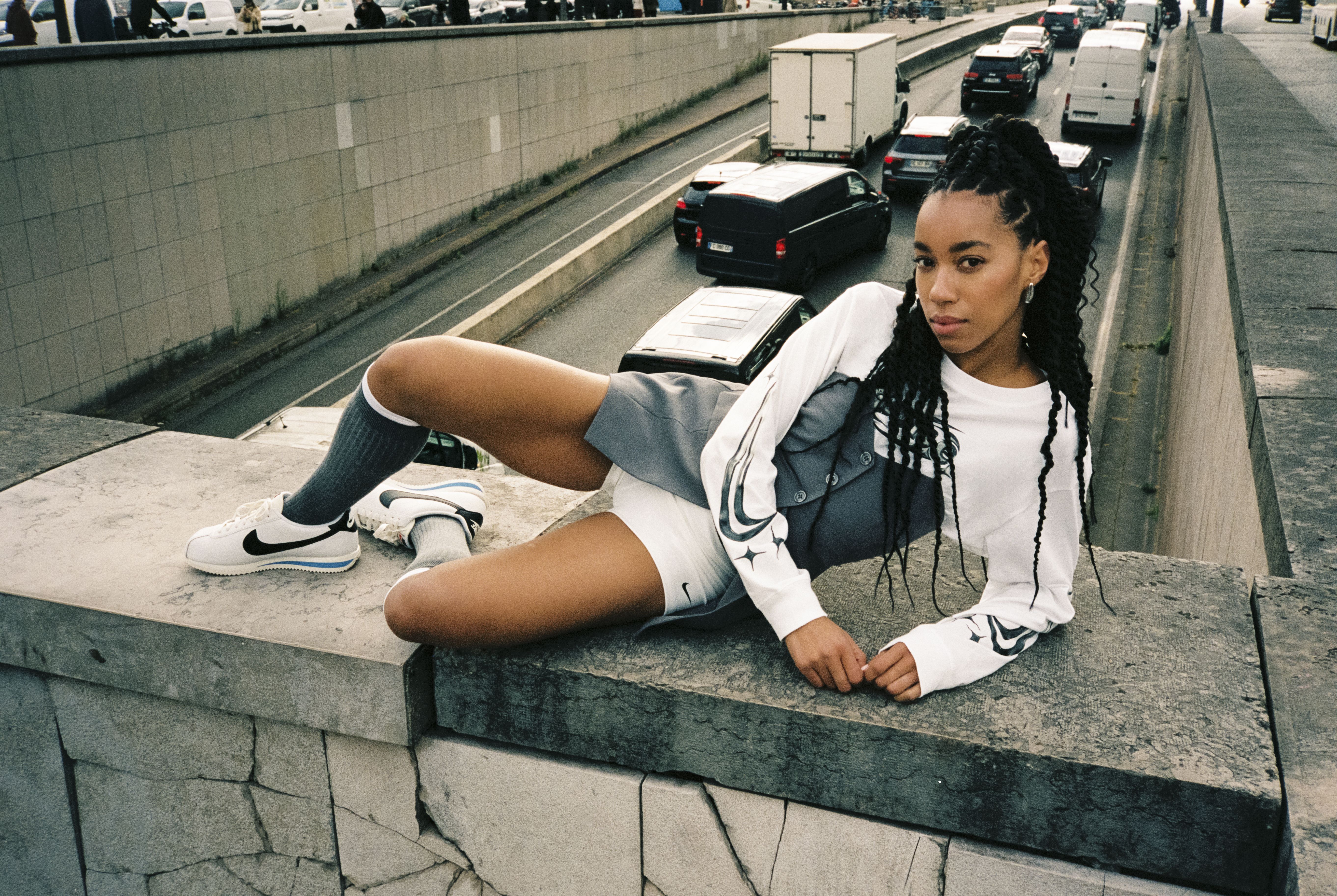 Žena u bijelom NIKE puloveru i bijelim NIKE tenisicama pozira na betonskom zidu iznad prometne ulice